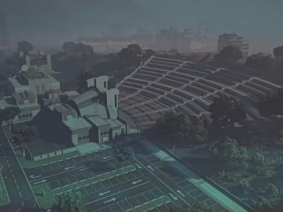 The image presents an aerial view of a complex in East Suffolk, United Kingdom. It features a cluster of modern, geometric buildings with flat roofs and a light-colored facade. One prominent structure is topped with a satellite dish. Adjacent to the main buildings is a large, sloping structure comprised of multiple parallel rows, resembling a stadium or greenhouse. A large parking lot with clearly marked spaces and directional arrows is visible in the foreground. A road runs alongside the parking lot and connects it to the complex. The scene is surrounded by trees and greenery, with some buildings visible in the distant background. The overall color palette is muted, with a predominance of gray and green hues. The scene appears to be shrouded in a light mist or fog, giving it a slightly hazy appearance.