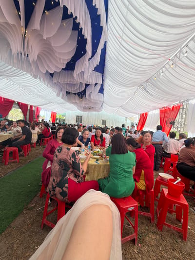 The image shows an outdoor event in Tay Son Commune, Vietnam. Many people are seated at tables under a large tent structure decorated with white fabric draping and blue sections. The tent also features some lighting elements. People are dressed in various clothing, suggesting a social gathering such as a wedding or a party. Red plastic stools are used as seating. The ground is a mixture of grass and bare earth.