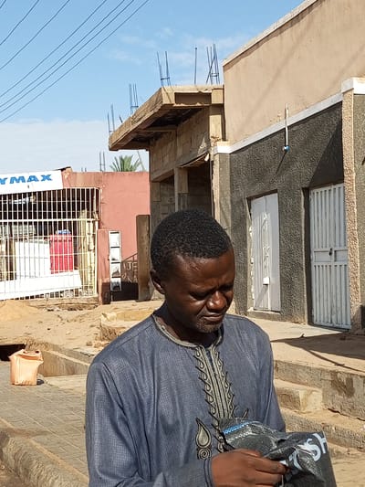 In Arawa, Nigeria, a man in traditional clothing is captured in a close-up shot. He wears a blue garment with intricate embroidery around the neckline and chest. His head is bowed, and he is holding a black bag with white lettering in his hands. In the background, a building under construction is visible, with exposed rebar and concrete. Adjacent to it, another building with a textured dark gray facade and white doors stands. The scene is set on a dusty street with a paved walkway, and a light-colored plastic bag is placed on the ground to the left. The sky above is clear and blue, with power lines crisscrossing it.
