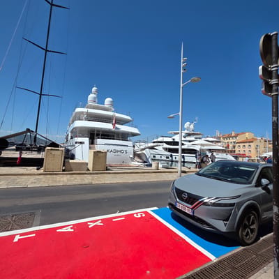 This image depicts a sunny day in a marina, likely in Saint-Tropez, France, given the location cue. The scene is dominated by several large white yachts docked along the waterfront, with a few people visible on and around them. In the foreground, a light gray Nissan car is parked on a designated area marked with red and blue paint. White lettering on the red section spells out "TAXI" vertically. The car has a French license plate reading "GV-898-VG". In the background, buildings with terracotta roofs are visible. A sailboat with black sails is also partially visible on the left side of the image. The sky is a clear, bright blue, indicating good weather. The overall atmosphere suggests a lively and upscale maritime setting.