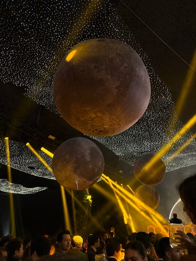 The image depicts an indoor event space with a dark ceiling adorned with numerous small, white lights resembling stars. Suspended from the ceiling are several large, spherical decorations designed to look like planets or moons, with textured surfaces and illuminated by yellow spotlights. These spotlights emanate from below, creating beams of light that cut through the smoky or hazy atmosphere.
Below the suspended decorations, a crowd of people is visible, with their faces and upper bodies in focus. They appear to be gathered for an event, possibly a party or celebration. Some individuals are looking towards the decorations, while others are engaged in conversation or holding drinks. The overall ambiance is festive and dimly lit, with the yellow lights and starry ceiling creating a dramatic effect.
The description of this image does not provide any information about the location in the city of Monterrey, Mexico.