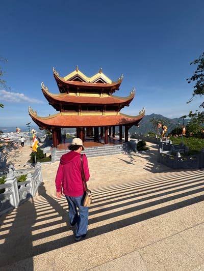 A daytime, eye-level, long shot captures a view outside a pagoda near Diên Khánh, Vietnam. The foreground consists of a person with their back toward the camera, wearing a red hooded sweatshirt, blue pants, gray shoes, and a baseball cap. They have a brown purse strapped to their side. The pagoda is to the left, showcasing red and gold paint, with a multi-tiered roof. Between the pagoda and the person is a balustrade, with flags in the distance. Beyond the balustrade, in the middle distance, is an overview of a city with some small mountains in the background. Above the pagoda and behind the mountains is a bright blue sky, slightly obscured by trees. Long shadows are cast across the brick walkway in the foreground.