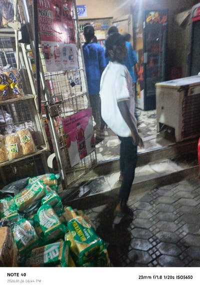 This evening image, taken on January 26, 2026, at 06:44 PM, captures a bustling scene at a local market stall or small shop in Panshekara, Nigeria. The lighting is dim, with artificial lights illuminating the interior of the shop and casting a glow on the immediate foreground.
In the lower left foreground, a large pile of packaged bread is prominently displayed, primarily featuring green and white wrappers. Visible text on some packages includes "MIJINYAWA Bakery" and "WHITE BREAD". To the left, a metal shelving unit holds more packaged bread, identifiable by its brown and gold wrappers, on its upper shelves.
Attached to the shelving unit are two promotional posters. The upper, larger poster is red and white, proclaiming "WIN N600M IN CASH PRIZES!" and details steps: "BUY SCAN PAY WIN", also mentioning "2. Milioni". Below it, a smaller pink poster advertises a beverage with the text "New Kook Same delicious taste!" alongside an image of a red bottled drink.
Several people are present in the scene. A person in a white t-shirt and dark pants stands in the mid-ground, with their back mostly to the viewer, their head turned slightly to the left. Further inside the shop, two other individuals are visible, less distinctly, both wearing blue shirts.
To the right in the background, a commercial refrigerator unit with an exposed condenser coil is discernible, indicating cold storage facilities. The ground is paved with patterned tiles or bricks, appearing somewhat reflective in areas, possibly from moisture or the overhead lighting. The overall ambiance suggests a busy local commercial hub during the evening hours. The image quality, as indicated by the EXIF data, was captured at 23mm, f/1.8, 1/20s, and ISO5650.