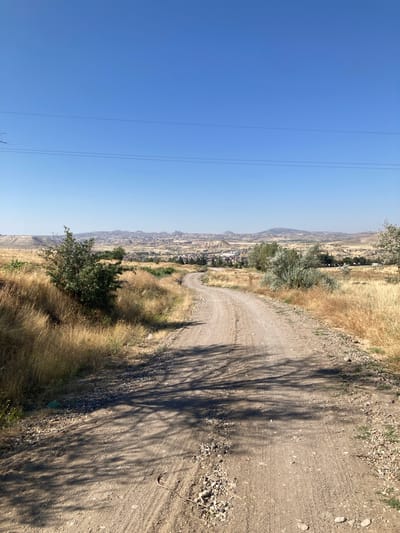 The image depicts a dirt road winding through a dry, grassy landscape under a clear blue sky. In the distance, the city of Avanos İlçe Merkezi, Turkey, is visible with its characteristic rock formations and buildings. The road shows tire tracks and is bordered by dry, yellow grass and scattered rocks. Sparse trees and bushes dot the landscape, casting shadows on the road. Power lines are visible in the upper part of the image, stretching across the sky. The scene appears to be a rural area on the outskirts of the city, bathed in bright sunlight, suggesting a warm, dry climate. There are no people or animals clearly visible in the image.