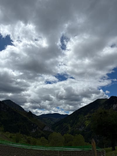 A vertical outdoor shot features two mountain ranges. The mountains are covered in lush green vegetation. Between them, the sky is visible. Most of the sky is obscured by thick, white clouds with patches of blue sky peeking through. The terrain at the foot of the mountains appears flat and green, potentially cultivated. A partially visible structure can be seen, constructed from green netting attached to white posts. The image's location is Giovren, Bulgaria.
