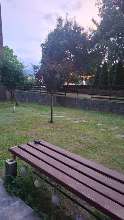 This is a vertical, eye-level, outdoor photograph of a backyard area in Razlog, Bulgaria, taken at dusk. In the foreground, a dark brown wooden bench with metal legs occupies the bottom right of the frame, facing left and slightly away from the viewer. A dark object, possibly a phone, rests on the bench. Hanging from the underside of the bench are three white spherical lanterns. To the left of the bench, a small solar-powered garden light is visible. The ground beneath the bench is covered in green grass and some leafy plants.
Behind the bench, a well-maintained lawn stretches back to a dark stone retaining wall. A dark wooden fence runs along the top of the retaining wall. Beyond the fence, several trees of varying sizes and types are visible, with some featuring small, glowing yellow lights strung between them. The sky above is a soft blend of pale blue and pink, indicating twilight. The overall impression is a peaceful, semi-urban backyard setting at the end of the day.