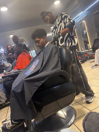 The image shows the interior of a barbershop. A Black male barber wearing a black and white patterned sweater, black gloves, and a black hat is cutting the hair of a Black male customer who is draped in a black barber cape. Other customers are visible in the background, seated and waiting their turn. The environment includes barber chairs, mirrors, and tiled floors. The storefront signage visible in the mirror indicates the location is in Blouberg, South Africa, identified by the telephone area code "061216".