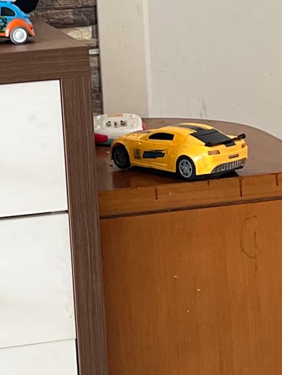 A yellow toy car is precariously balanced on the edge of a wooden surface. The car is a sports car model, with black accents and detailing. It appears to be a die-cast model, possibly a replica of a performance vehicle. The wooden surface is a rich brown color and has a textured grain. Several small, dark marks are visible on the wood, possibly from wear and tear or age. To the left of the car, a small white electronic device with buttons and a screen is partially visible. Further to the left, part of a blue and orange toy vehicle is visible, with a white wheel and orange accents. The background on the left is a textured wall, likely in a home setting. The overall impression is of a child's play area or a shelf where toys are stored. There is no discernible activity taking place, as the car is stationary. The lighting suggests an indoor environment, possibly during the daytime. There is no visible text on the toy car that can be read clearly.