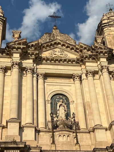 This is a vertical, eye-level, close-up shot of the facade of a grand, ornate building, likely a church, in Catania, Italy. The architecture is Baroque, characterized by elaborate carvings, columns, and decorative elements. The stone material of the building appears to be light-colored, possibly sandstone or limestone, and is illuminated by bright sunlight, casting distinct shadows.
The primary subject is the architectural detail of the building. There are several large, fluted columns adorning the facade, topped with intricate Corinthian capitals. Between the columns are carved friezes and pediments, featuring figurative sculptures and geometric patterns. In one prominent section, a semicircular arched window is visible, containing a stained-glass depiction of what appears to be a religious scene with figures. A statue of a robed figure, possibly a saint, is positioned on a small balcony above this window, accompanied by another smaller figure. A wrought-iron railing and a decorative lamppost are also present on this balcony.
At the very top of the visible structure, a cross with a distinctive pointed finial stands against a backdrop of a bright blue sky dotted with fluffy white clouds. This suggests it is daytime, likely late morning or early afternoon, given the strong sunlight and clear skies. The clouds indicate fair weather. The overall impression is one of grandeur, history, and religious significance. No text is visible in the image.