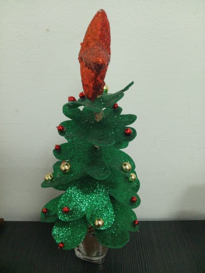 A close-up, medium shot captures a miniature, glitter-covered Christmas tree, its green felt branches adorned with small red and gold spherical ornaments. Atop the tree sits a cone-shaped topper, also glittery and red. The tree is positioned on a dark, textured surface, with a white, textured wall as the backdrop. The lighting is even, suggesting an indoor setting. There are no people visible in the image. This image does not provide any information that would allow for a determination of its location within Guayana City, Venezuela.