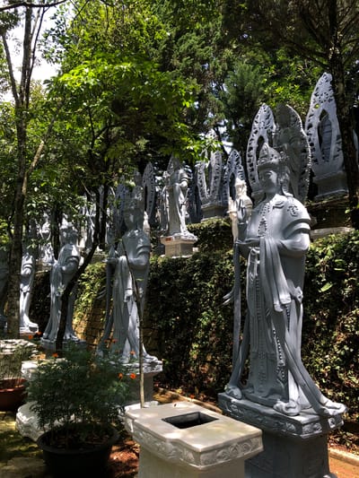 The image shows a series of grey, stone statues of human figures arranged in a line, likely part of a religious or decorative display. The statues appear to be of women, dressed in ornate traditional clothing with elaborate headwear. They are positioned outdoors in what looks like a garden or park setting, with lush green trees and foliage surrounding them. Sunlight filters through the leaves, casting shadows on the statues and the ground, suggesting it is daytime. The statues are weathered, indicating they may be old or have been exposed to the elements. The scene is peaceful and serene, with a sense of stillness and reverence. There are no visible text elements in the image. The presence of statues and the natural surroundings suggest a location of cultural or spiritual significance, possibly a temple, a memorial, or a garden attraction. Given the location context of Nam Ban, Vietnam, these statues could be representations of deities, historical figures, or elements of local folklore.