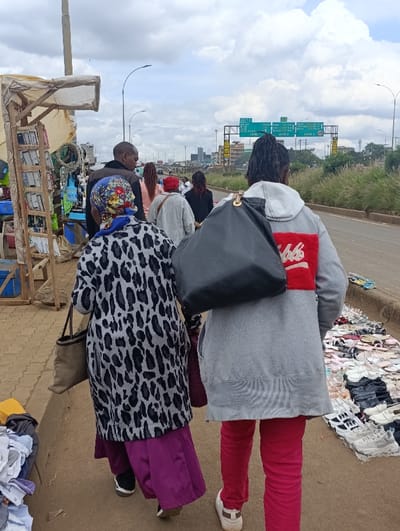 The image shows a street scene in Githurai, Kenya. The scene is set on a paved road with a sidewalk. A group of people are walking along the sidewalk. Two women are in the foreground, a woman in a grey and black leopard print coat with a purple skirt and a brown bag in her left hand, and a woman in a grey sweatshirt with red pants and a black bag slung over her right shoulder. The woman in the leopard print coat is walking ahead of the woman in the grey sweatshirt. Behind them is a man in a blue shirt. The woman in the grey sweatshirt is looking at the camera. There is a street vendor stall on the left side of the image with items for sale, including books and other goods. There is a sign on the right side of the image indicating the exit of the highway with lanes marked for various destinations. In the background are buildings and vehicles. The scene is busy with people and vehicles.
