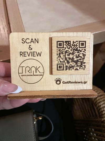 A close-up, slightly tilted shot captures a person's hand holding a small, light-colored wooden stand. The hand features neat nails with a French manicure. The wooden stand is the primary subject, designed to encourage customer engagement.
Dominating the upper section of the stand is a laser-engraved QR code, positioned on a slightly raised square piece of wood. To the right of the QR code, the text "GetReviews.gr" is visible, accompanied by a small icon depicting a thumbs-up hand surrounded by three stars, suggesting a review platform.
Below the QR code, the words "SCAN & REVIEW" are engraved vertically. Further down, a circular logo encloses the brand name "TRNK", with "COFFEE | COCKTAIL | BRUNCH | DRINKS" written beneath it, indicating the services offered by the establishment.
The background is softly blurred, showing a portion of a dark woven material, possibly a basket or furniture, and a segment of a beige handbag. The overall scene suggests an indoor setting, likely a cafe, bar, or restaurant in Chaidari, Greece, where the stand serves to prompt customers to leave online reviews.