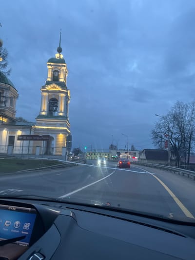 The image, taken from inside a car, shows a view of a road and a church in the city of Ново-Никольское, Russia. The road, marked with yellow and white lines, stretches ahead, with cars visible in the distance. To the left of the road, a cream-colored building with a green dome and a tall, slender bell tower with a clock on its face stands prominently. The sky above is a muted blue-gray. The car's dashboard is visible in the foreground, showing a digital clock displaying "04:36".