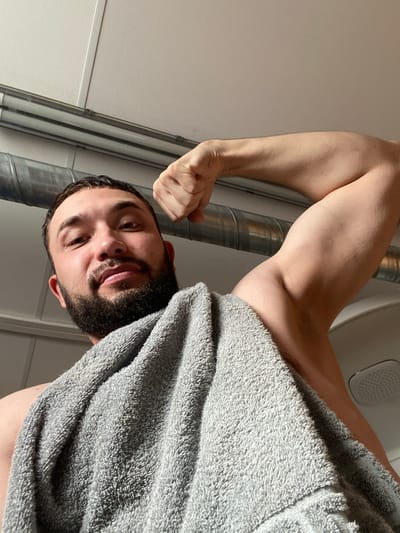 A man with dark hair and a beard is looking down at the camera. He has a gray towel draped over his shoulder and is flexing his bicep. The background appears to be a bathroom with metal pipes and a white wall. The location is not determinable from the image.