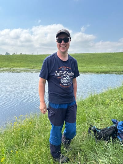 A man is standing on the grassy bank of a body of water, likely a river, with a green embankment behind him under a partly cloudy sky. He is wearing a dark blue t-shirt with a graphic print that reads "Taste of Freedom" and features a motorcycle, sunglasses, and a dark baseball cap. His pants are a combination of dark blue and lighter blue panels. To his right, on the grass, are a black fanny pack and a blue backpack. The overall setting appears to be a rural or park area, possibly in the village of Sredneye Kechevo, Russia, given the description.