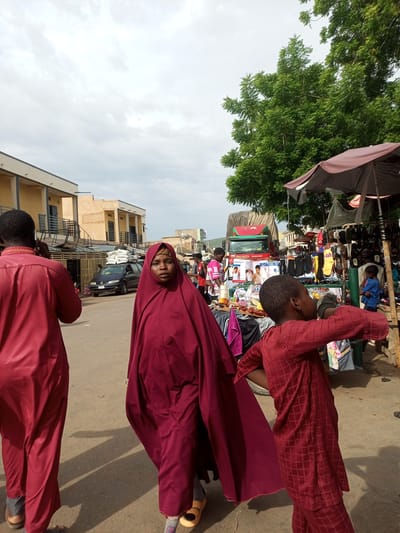 The image depicts an outdoor market scene in Arawa, Nigeria. In the foreground, a woman wearing a dark red hijab and a matching flowing garment walks toward the camera. To her left, partially obscured, is a person in a matching red outfit. To the right of the woman, a young child in a red patterned garment is walking away from the camera. In the background, a dark blue van is parked on a dirt road, laden with white sacks on its roof. Beyond the van, stalls are set up with various goods for sale, including clothing and footwear, beneath a large green tree. Buildings with light-colored facades and balconies line the left side of the street. A large red truck is visible further down the road. The sky is overcast with scattered clouds.