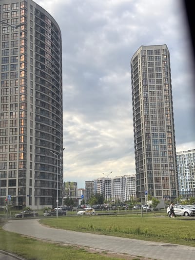 The image depicts a modern urban environment, likely in the Сеніцкі сельскі Савет, Belarus, characterized by towering residential buildings. In the foreground, a curved sidewalk runs alongside a grassy area. Beyond the grass, a street is visible with several cars parked and in motion. Two prominent, tall, curved buildings dominate the mid-ground, featuring numerous windows and balconies, suggesting apartment complexes. Further in the distance, more buildings of varying heights and architectural styles are present. The sky is overcast with clouds. On the right side of the image, two people are walking on the grass next to parked cars. One of the cars is white, and another is silver. A street sign with a parking symbol is also visible.