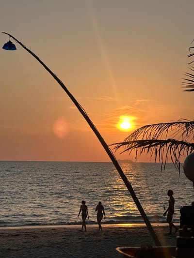 This image captures a sunset over a beach in Phra Ae, Thailand. The horizon shows a setting sun, creating a bright orange and yellow sky with a reflective path on the ocean surface. The ocean exhibits calm conditions with small waves reaching the sandy beach. A tall, dark pole with a blue, downward-facing light fixture at its apex extends diagonally from the lower left towards the upper right. Silhouetted palm fronds are visible on the right side of the frame. Three human figures are silhouetted on the sandy beach near the waterline. Two figures, presumed female and wearing swimwear, walk from left to right. A third figure, presumed male and wearing swimwear, walks from right to left, positioned further right. A faint, dark outline consistent with a distant island or large vessel is present on the horizon, directly in line with the sun's reflection. A partially visible, blurry red and yellow object, possibly a container, occupies the extreme lower right foreground.