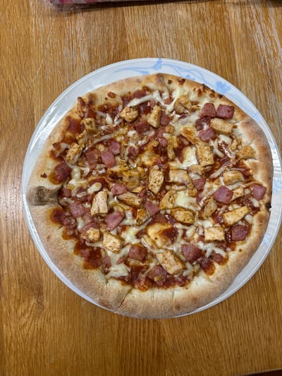 A top-down view reveals a pizza placed on a white plate with a delicate blue floral pattern, resting on a wooden table. The pizza is topped with chunks of seasoned chicken, diced ham, and melted cheese over a tomato-based sauce. The crust is golden brown and appears homemade. The setting seems to be indoors, likely a home dining area, but the specific location in Oswestry, United Kingdom, cannot be determined from this image alone.