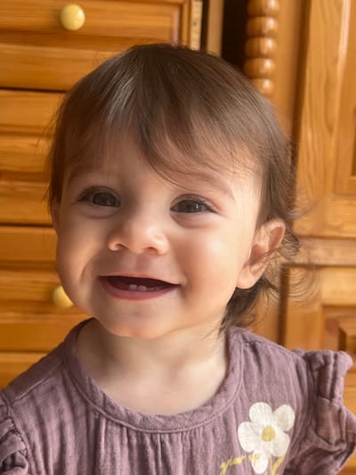 A close-up, head-and-shoulders shot shows a smiling baby girl in the foreground. She has dark, wavy hair that falls to her shoulders and is looking directly at the camera. Her eyes are dark, and her mouth is open in a wide smile, revealing a few small teeth. She is wearing a light purple shirt with ruffled sleeves and a white flower with a yellow center appliqued on the left side of the chest. The background is a wooden dresser with several drawers. A yellow drawer knob is visible on the top left of the frame. The lighting is warm and natural, suggesting the photo was taken indoors during the day. The location of this image is not specified, but it is described as being in the city of Elena, Bulgaria.