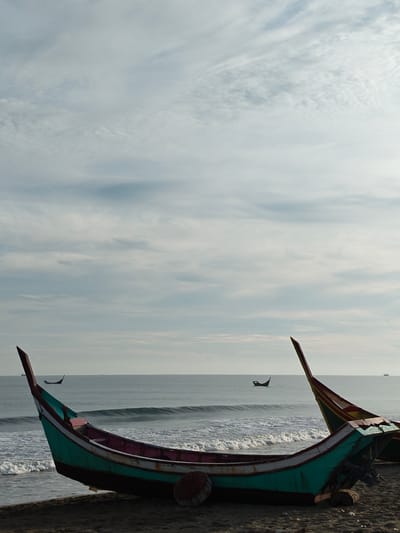An overcast sky with wispy clouds stretches above a serene ocean and a dark sandy beach in Curehtunong, Indonesia. The foreground is dominated by the hull of a traditional Indonesian fishing boat, its vibrant teal and pink colors contrasting with the muted tones of the surroundings. The boat rests on the sand, partially submerged in the incoming tide, with gentle waves lapping at its sides. In the distance, two other smaller fishing boats are visible on the calm water, appearing as dark silhouettes against the hazy horizon. The overall atmosphere is one of quiet solitude and the gentle rhythm of coastal life.