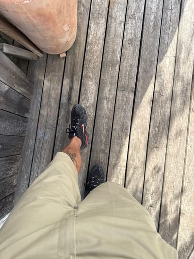 The image shows a person's legs and feet on a weathered wooden deck. The person is wearing tan pants and black athletic shoes with red accents. One shoe is in focus, revealing a portion of the person's tattooed leg. To the left, a large, round terracotta object rests on the deck. The wooden planks are grey and show signs of age, with visible grain and knots. Sunlight casts shadows across the deck, suggesting it is daytime. The overall impression is casual and outdoorsy, possibly a backyard or patio setting in Vilanova i la Geltrú, Spain. No text is visible in the image.