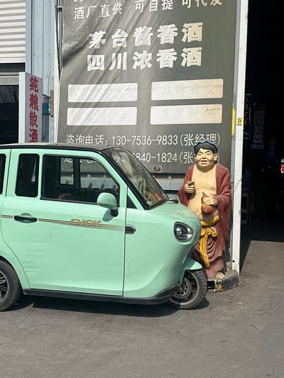 A mint-green three-wheeled electric vehicle, marked "CHOK" on its side, is parked on a paved surface in Licheng District, China. To the vehicle's right stands a colorful statue of a laughing Buddha figure, dressed in a red robe and yellow sash, holding a reddish-brown gourd. Behind the vehicle and statue, a large dark banner affixed to a building advertises "Maotai Jiangxiang Wine" and "Sichuan Nongxiang Wine" in white Chinese characters, including consultation phone numbers for "Manager Zhang." To the left, a vertical red sign displays "Pure Grain Bulk Liquor" in white Chinese characters. The scene is illuminated by daylight, suggesting an outdoor commercial setting.