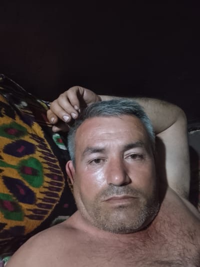 A man is lying down. He has gray hair, a beard, and is shirtless. His right arm is raised and bent behind his head. He is lying on what appears to be a patterned blanket or cushion. The background is dark, possibly a wall or ceiling. There is no information about location in Sabir Rakhimov, Uzbekistan.