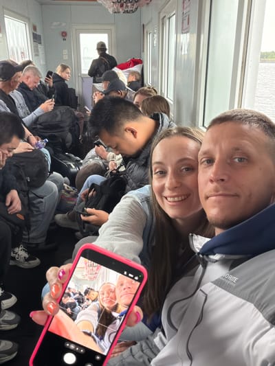 A man and a woman are captured in a selfie taken from a first-person perspective, with the woman holding a bright pink smartphone that displays the live camera feed. The couple is positioned head-to-head, both smiling at the camera. The man wears a grey and navy jacket with the text "FAVORITE DESIGNATED IN 1993" visible on the sleeve. The woman has long, dark hair.

The scene unfolds inside a crowded cabin, likely a ferry or train, given the configuration of windows, seating, and overhead structures. Numerous other passengers are visible in the background, many with backpacks and luggage, suggesting they are in transit. Most passengers appear to be seated, some looking at their phones, others resting.

Daylight filters through the multiple windows along the side of the cabin, indicating it is daytime, though the exterior view suggests an overcast sky. The overall atmosphere points to a shared travel experience. The specific location, Nizhneleninskoe, Russia, reinforces the context of a journey, possibly involving a border crossing or ferry service. The phone's screen shows typical camera app interface elements including a shutter button, grid lines, and mode selection icons.