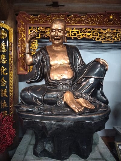 A statue of a man, likely a Buddha figure, sits cross-legged on a pedestal. The statue is composed of a dark metal material that contrasts with the gold coloring of his skin. The man is depicted wearing a flowing robe. His right hand is raised and holds a small object between his fingers. He has a bald head and an open mouth. The statue is set against a backdrop of ornate gold and dark wood paneling. The sign on the left side has Chinese characters written vertically in gold lettering. A bouquet of red berries or flowers is visible near the left side of the base. The location is Hong Gai Ward, Vietnam.