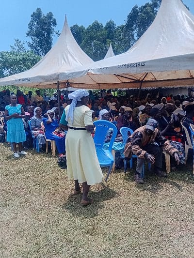 This image displays an outdoor gathering taking place under two white, conical-roofed event tents on a sunny day in Butali Chegulo ward, Kenya. A large group of individuals, predominantly adults, are seated on a variety of chairs, including numerous blue plastic chairs, beneath the tents. The underside of one tent displays partial text, including the sequence "0748821915." In the left foreground, a woman wearing a light blue dress and white sneakers stands facing right. Centrally, a woman in a light cream-colored long-sleeved dress with a black belt and a white headscarf stands with her back partially turned to the camera, also facing right. In the right foreground, a man is seated in a blue plastic chair, leaning forward with his head down, dressed in a brown patterned jacket and dark trousers. The ground consists of dry, yellowish-green grass. The background features lush green trees under a clear blue sky.