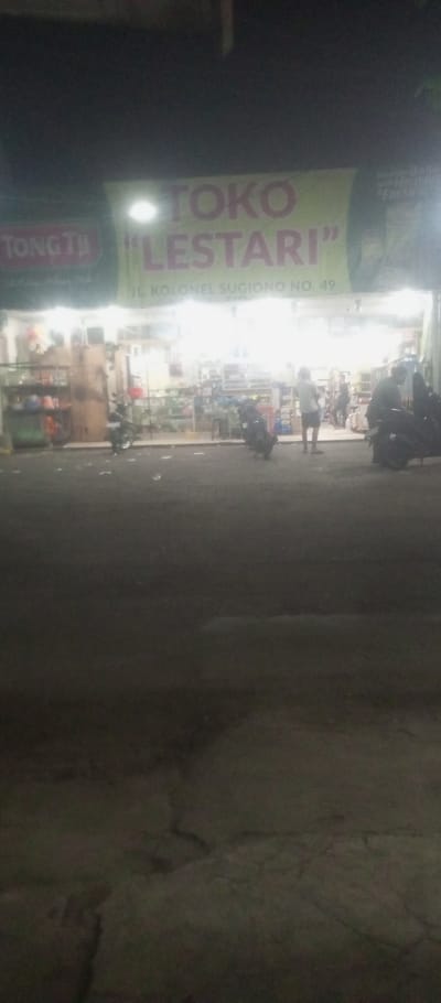 The image depicts a nighttime scene of a brightly illuminated retail establishment located at Jalan Kolonel Sugiono No. 49 in Kepuh Kiriman, Indonesia. The store is identified by a large sign displaying "TOKO LESTARI" in prominent pink letters on a light green banner, with "TONG TA" also visible on a green sign to the left. The store interior is highly lit by artificial light, revealing shelves stocked with various goods. At least three individuals are visible within or immediately outside the store's entrance; one person, wearing a white short-sleeved shirt and dark shorts, stands near the entrance facing inward. Two motorcycles are parked on the paved area in front of the store, facing the street. The foreground consists of a dark, paved road, which appears to have some scattered debris near the storefront. The overall environment is dark, indicating a nocturnal setting.