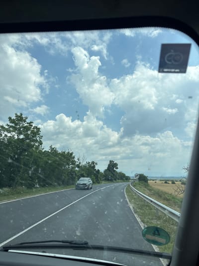 The image captures a view from a vehicle's windshield as it travels along a paved road. The road curves to the right, bordered by a grassy shoulder and a metal guardrail on the right. To the left of the road, dense green trees and foliage line the landscape. In the distance, a field of dry, yellowish vegetation stretches towards a hazy horizon. The sky is a bright blue, punctuated by numerous fluffy white clouds. A dark-colored car is visible driving ahead on the road, and a circular green sticker with a symbol is affixed to the lower right portion of the windshield. A square sticker with a circular emblem is also present on the upper right of the windshield. The scene appears to be in Osetenovo, Bulgaria.