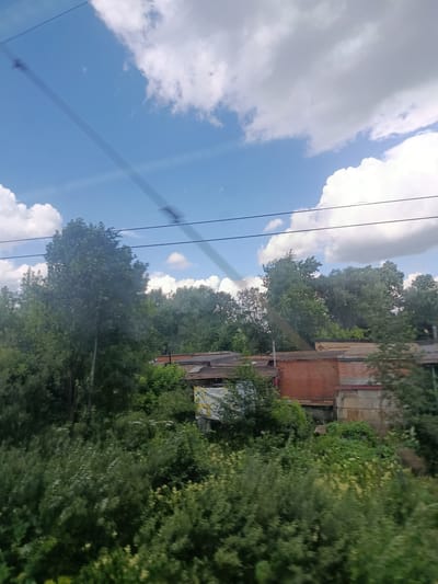 The image, taken from a moving vehicle, captures a view of a suburban or semi-rural area in Березка-291 Снт, Russia. The scene is dominated by lush green foliage, with several large trees in the foreground and middle ground. Behind the trees, the roofs of several buildings are visible, including a structure with red brick walls and a lighter-colored wall with visible concrete blocks. The buildings appear to be single-story structures, possibly garages or small outbuildings. The sky above is a vibrant blue, dotted with fluffy white clouds. Overhead, dark power lines are visible, running diagonally across the frame, suggesting the presence of infrastructure in the area. There are no people or animals visible in the image. The overall impression is one of a somewhat overgrown, natural environment interspersed with modest man-made structures.