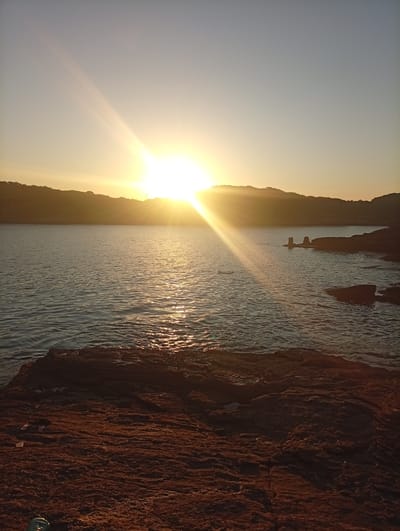 The image shows a sunset over the ocean in Armação dos Búzios, Brazil. The sun is setting behind a hill in the distance, casting a golden light over the water. The foreground is a rocky shore. There are no people visible in the image. Two cement pillars protrude from the water. Debris is on the rocks in the lower left corner.