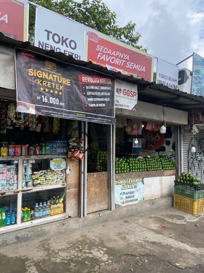 The scene is a small storefront in Lembang, Indonesia, located at the coordinates -6.817 latitude and 107.613 longitude. The store is a simple structure with a wooden frame and a corrugated metal roof. The storefront is covered in signs in Indonesian and English. One sign advertises "Signature Kretek" cigarettes for 16,000 Indonesian Rupiah. Another sign advertises "Revolusi Kretek," which appears to be a brand of cigarettes. The store window is filled with packaged goods, including drinks, snacks, and tobacco products. The store is open and there is a small amount of product displayed inside and outside the store. There are stacks of avocados in the corner, and a sign in Indonesian says "DISINI BISA BIKIN JUS ALPUKAT!!" which translates to "Here you can make avocado juice!". There is a stack of green avocados behind the store window. The storefront is located on a busy street with a concrete sidewalk and asphalt street. There are other buildings and structures visible in the background. The weather is overcast and grey. There are no people in the photo.