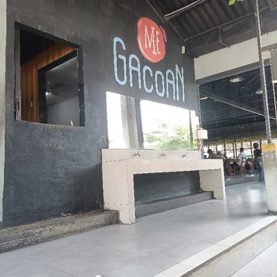 The image shows the exterior of a building in Kecamatan Ngaliyan, Indonesia (latitude: -7.002, longitude: 110.342). The building's facade is mostly black and features a large, blue sign that reads "ME GACOA[N]" with the word "ME" in a red circle. There is a window to the left of the sign and a large mirror extending to the right. In front of the building is a white, rectangular sink with three faucets. There is a step leading up to the sink. The sink is partially obscured by a large step leading up to a platform that appears to be part of the restaurant. There are several people sitting and standing at tables in the background. The tables are made of wood and have metal chairs.  The area is lit by overhead lighting and the floor is tiled.  The scene is empty except for the people in the background. The building is a restaurant or food stall and it appears to be open.