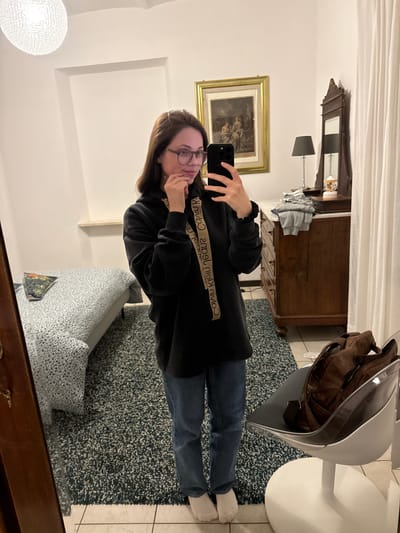 A woman is taking a selfie in front of a mirror in an interior setting. The woman has long brown hair, wears glasses, a black sweatshirt with "Calvin Klein Jeans" printed on it, and blue jeans. She is wearing white socks. The room features a wooden dresser with a framed painting hanging above it, a white ceiling lamp, a blue and white patterned armchair, and a gray chair with a brown backpack on it. A blue and gray shaggy rug covers the floor. The location is likely in Perugia, Italy, as the prompt specifies this location.