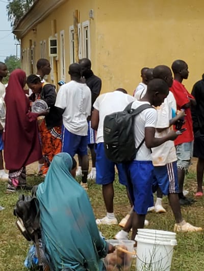 In Bahit, Nigeria, a group of young people are gathered near a yellow building. The building has white-trimmed windows and appears to be a school or community center. Several individuals are wearing casual clothing, including t-shirts, shorts, and head coverings. One person in the foreground is wearing a teal-colored hijab and a dark bag rests on their lap. Another person, wearing a maroon hijab, is holding a silver bowl. The ground is covered in grass and some scattered debris. A white bucket and a clear plastic container with what appears to be food are also visible on the ground. The overall impression is of a casual gathering, possibly outdoors for an event or meal.