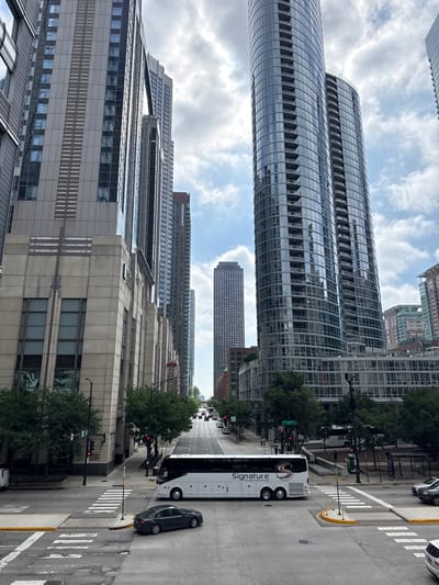 The image captures a street-level view of a city intersection in Chicago, Illinois, USA, characterized by towering skyscrapers and urban infrastructure. In the foreground, a white tour bus with "Signature Transportation Group" printed on its side is positioned in the middle of the intersection, having just passed a crosswalk. A dark sedan is in the process of turning, also having crossed a crosswalk. The intersection is marked with white lines and yellow bollards. Traffic signals are visible, with one showing a red light.
The environment is dominated by modern high-rise buildings. To the left, a light-colored building with large windows and vertical detailing rises high, featuring a sign with what appears to be artwork. Further down the street, another tall building with a textured facade is visible. On the right side of the image, two prominent, curved skyscrapers with numerous balconies and reflective glass facades dominate the skyline. These buildings are interspersed with smaller buildings and trees lining the sidewalks.
In the background, more tall buildings stretch towards the sky, and a multi-lane street with several vehicles can be seen moving away from the viewer. The sky is partly cloudy, with patches of blue sky visible through the white clouds. The overall impression is a dynamic urban scene in a bustling metropolitan area.
