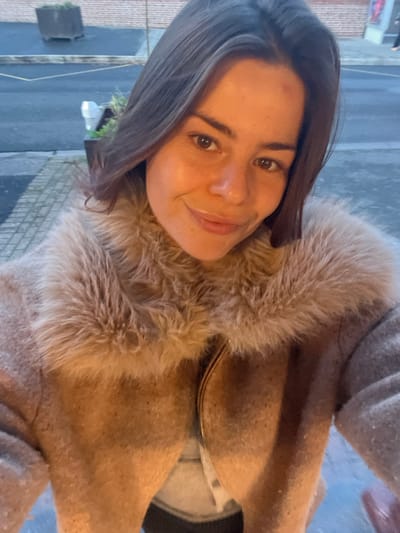 A single female individual is depicted in a close-up, high-angle selfie taken outdoors in Villeneuve-d'Ascq, France. She possesses dark brown hair and fair skin, with a visible light mole on her forehead, and is looking directly at the camera with a slight smile. She wears a tan or light brown jacket featuring a prominent, fluffy faux fur collar in a lighter beige hue. A golden metallic zipper is partially visible down the front of the jacket, with a lighter, possibly grey, garment visible underneath. The background shows an urban environment at what appears to be dusk or dawn, characterized by dim, ambient lighting. A dark asphalt road with faint yellow markings occupies the lower background, adjacent to a light-colored paved sidewalk. On the sidewalk, two dark, square-shaped planters containing green foliage are positioned. Behind these, a reddish-brown brick wall is partially visible, indicating a building facade. No other persons or significant actions are present in the visible scene.