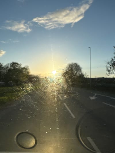 The image shows a landscape at sunset through a dirty window. The sun is setting over trees, with a blue sky and clouds visible. A streetlight is present in the midground. On the right, the window's interior is visible, showing a black dashboard with round details and faint markings. The image appears to be taken from inside a vehicle. Oswestry, United Kingdom, is not discernible from this image.