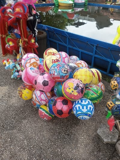 A collection of colorful inflatable balls are stacked on a textured surface. The balls feature soccer ball patterns, cartoon characters, and various designs. To the left, traditional lion dance puppets and toys are visible. In the background, a blue-lined pool or tank holds small paddle boats. The scene appears to be an outdoor market or entertainment area.