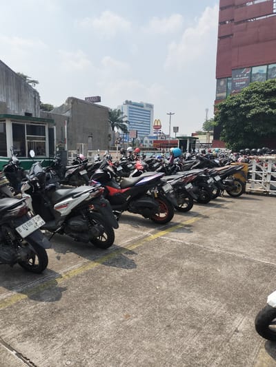 The image shows a parked motorcycle on a concrete surface with yellow lines. In the background, there are several other motorcycles parked in rows, along with a building that has a sign reading "MAKO". Further in the background, there are tall buildings and a McDonald's sign. The scene appears to be an outdoor parking lot in a city, possibly in Bekasi, Indonesia, given the presence of Indonesian license plates on some vehicles and the architectural style of the buildings. The sky is visible and appears to be partly cloudy.