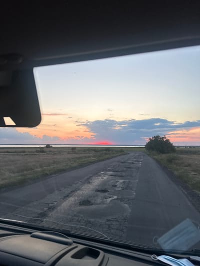 This image was taken from inside a vehicle, looking forward through the windshield. The scene depicts a sunset over a body of water. A cracked and potholed asphalt road stretches into the distance, flanked by dry, grassy fields. In the foreground, a portion of the vehicle's dashboard is visible, along with a light blue face mask lying on the windshield. The sky is painted with the warm hues of sunset, with soft clouds and a distinct band of red light low on the horizon, indicating the sun is either rising or setting over the water. A solitary tree stands on the right side of the road, adding a focal point to the landscape. The overall impression is one of a rural setting at dusk, on a road in need of repair, with the natural beauty of the sunset dominating the background.