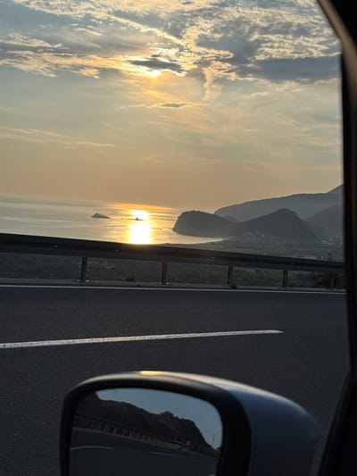 This is a vertical, eye-level, medium shot of a sunset over the sea, taken from a moving vehicle on a road in Kanjoši, Montenegro. The sun is low on the horizon, casting a bright, golden reflection on the water. In the distance, there is a small island. The sea meets the land, where rolling hills and mountains are silhouetted against the warm sky. A highway guardrail runs horizontally across the middle of the frame, and the asphalt road is visible in the foreground, with a white line marking the lane. A side-view mirror of the vehicle is in the lower right corner, reflecting the scene behind. The sky is filled with scattered clouds, some tinged with the colors of the sunset. There are no people or animals visible in the image.