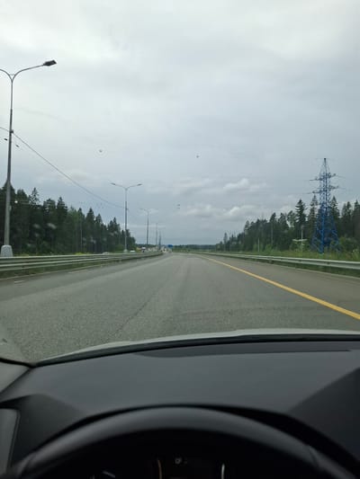 The image depicts a view from inside a vehicle traveling on a highway. The sky is overcast with gray clouds. On the left side of the road, there are streetlights and a dense forest. On the right side, there is a blue electrical tower and more trees. The highway has two lanes in each direction, separated by a yellow line. The road appears wet, suggesting recent rain. The foreground shows the dashboard and steering wheel of the car. This scene is likely on a highway near Lobnya, Russia, given the presence of typical Russian highway infrastructure. There are no people visible in the image.