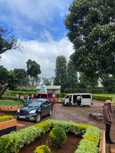 The image depicts an outdoor scene at Karatina Stage, Karatina, Kenya, under a partially cloudy sky. The ground is unpaved, consisting of damp, dark earth. In the immediate foreground, raised planters with green and white variegated hedges are present, bordered by low walls painted with orange and black stripes.
Three vehicles are prominent:
1. A dark-colored Honda CR-V SUV (license plate KCH 486H) is parked in the mid-ground on the left, facing right.
2. Behind the Honda, a red Nissan van or minibus, marked with a white stripe and a circular emblem, is also parked.
3. To the right, a white Toyota Hiace van or minibus is stationary with its passenger-side sliding door open. The front driver's side wheel appears to be detached or heavily damaged, with the vehicle resting on its hub.
Several individuals are present:
* In the foreground on the right, a male wearing a brown hooded sweatshirt, dark trousers, and a wide-brimmed straw hat stands facing left.
* Near the open white van, two individuals are visible; one in a light-colored top stands beside the open door, while another in a yellow top is partially visible inside the vehicle.
* A group of approximately 5-7 individuals, some appearing to be conversing, are gathered near the red van and behind the Honda CR-V on the left. One person in this group wears a yellow high-visibility vest.
In the background, a distinctive building with a multi-colored, tiered roof structure (featuring red, blue, and white sections) is visible, identifying it as part of the Karatina Stage. Tall trees and various green foliage frame the scene, with a concrete wall running along the mid-ground. A blue pole stands vertically between the red van and distant trees.