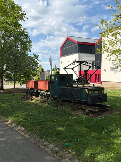 This image, taken in Pétange, Luxembourg, depicts a static display of an industrial locomotive and a small wagon set on tracks, situated in a grassy area next to a modern building.
The central subject is a dark green locomotive, number "2" painted in yellow on its side, featuring an intricate black pantograph-like structure on its roof. Attached to it is a rust-red, open-top wagon. Behind the wagon, a dark grey or black rectangular sign or marker is visible, with faint white text that appears to include "1940-19". The locomotive and wagon are positioned on a short section of railway track, which is embedded in well-maintained green grass. The grass is dotted with numerous small white and yellow flowers, resembling daisies and dandelions.
Adjacent to the grassy display area, a paved pathway runs along the right side of the frame. In the background, a striking modern building with a multi-layered design dominates. It features white walls, dark grey or black slatted panels, and prominent red structural or decorative elements, particularly on its roofline and around some window areas. Trees with vibrant green leaves frame the scene from the foreground and background, suggesting a spring or early summer setting.
The sky is bright blue with scattered fluffy white clouds, indicating clear and sunny weather. The overall impression is of a historical railway exhibit placed in a contemporary public space or campus-like environment. No people are visible, and there is no active event taking place, suggesting the scene captures a tranquil moment at a permanent outdoor display.